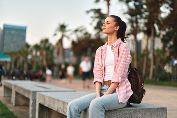 Fototapeta premium Woman in casual outfit sits on a park bench, backpack beside her, looking upward, peaceful outdoor scene with soft city backdrop