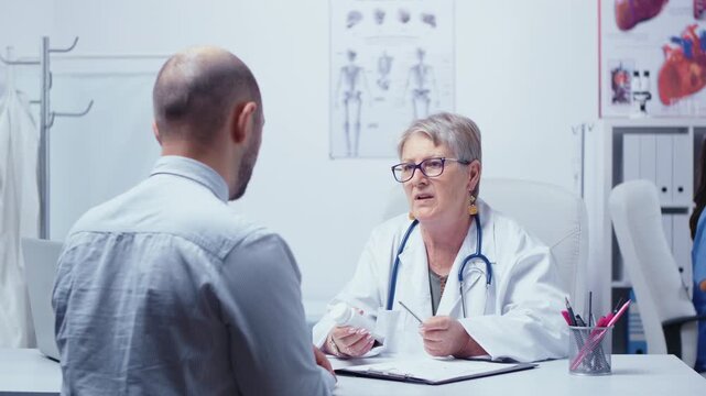 Experienced senior woman doctor talks about pills dosage with a male patient, guiding medication use during a private clinic consultation.