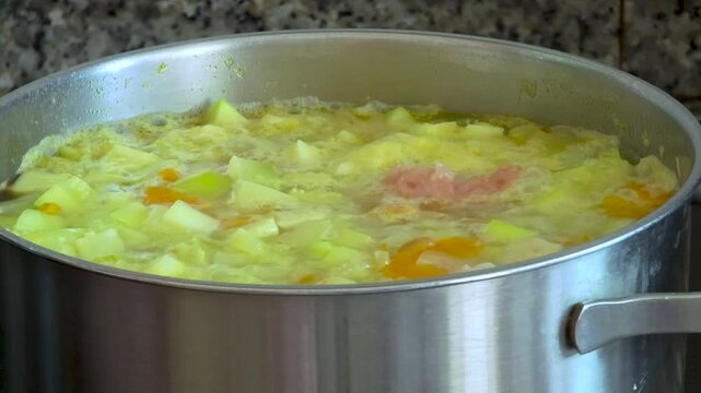 Slow motion close-up of adding raw minced meat dumplings into a boiling homemade vegetable soup with carrots and fresh dill in a stainless steel pot.