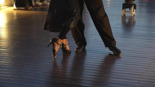Low angle detail of a man and woman's feet performing an argentine tango. The couple moves with passion on a wooden dance floor