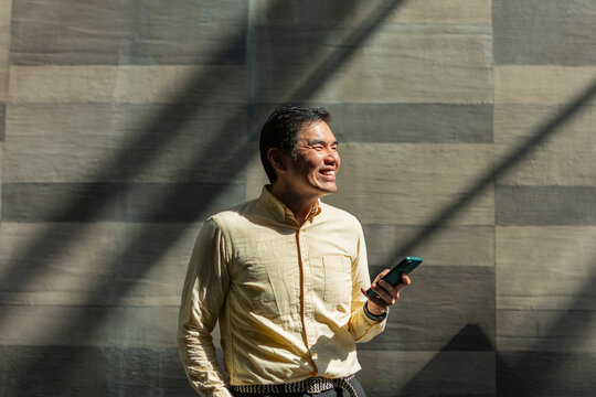 Smiling Asian man using smartphone in sunlit corridor