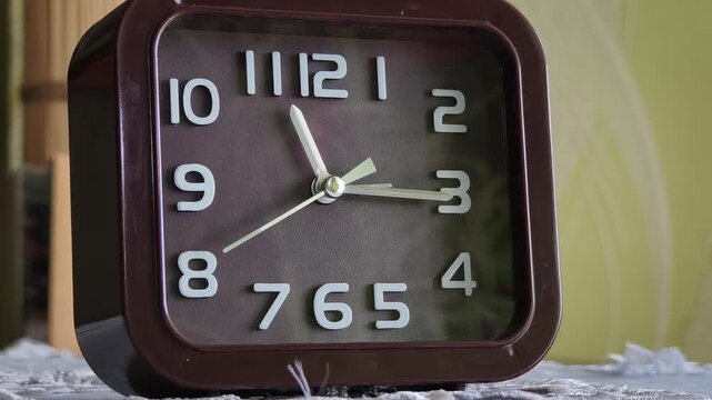 Close-up of a square brown analog alarm clock on a table with a sweeping white second hand. The sharp focus on the classic dial highlights the concept of punctuality.