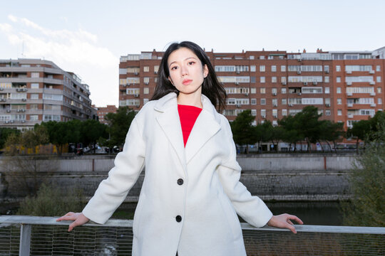 Gen Z Asian woman in white coat on city bridge at dusk