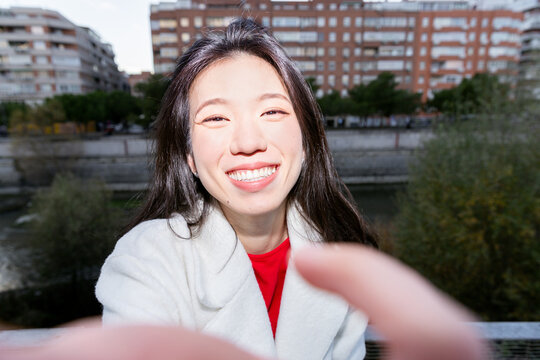 Smiling Gen Z Asian woman taking selfie on city bridge