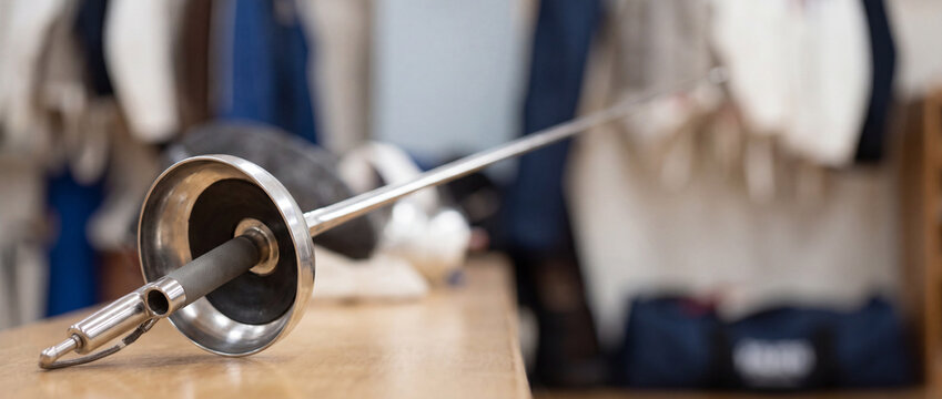Fencing epee lying on wooden bench in locker room with protective gear and jackets