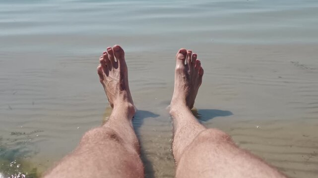 Point of view shot of a man's hairy legs and feet resting in the calm shallow water of a lake or sea. The camera slowly pulls back creating a sense of peace and relaxation on a sunny holiday.