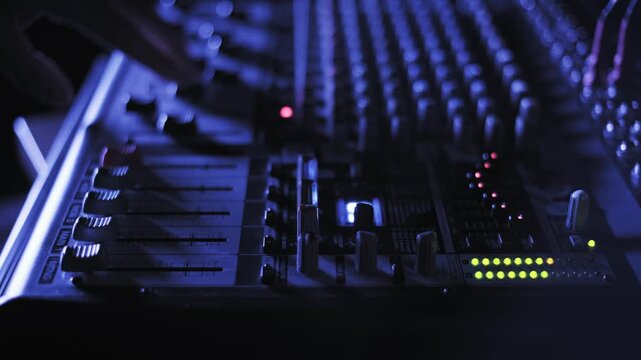 Hand of a sound engineer adjusts faders on a professional audio mixing console illuminated by blue light. The concept represents live music production and technical audio control.