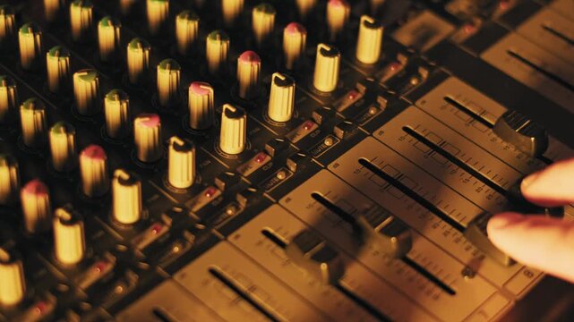 Close-up of a sound engineer's hand adjusting faders and knobs on a mixing console. Glowing buttons and level sliders are visible under dynamic blue and purple club lighting.