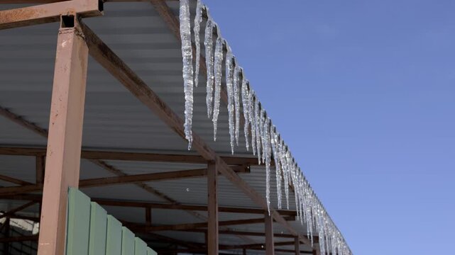 Sharp frozen ice spikes hang from a rusty metal roof structure and drip water against a clear blue sky. Concept of thawing temperatures, seasonal changes, and dangerous winter hazards.