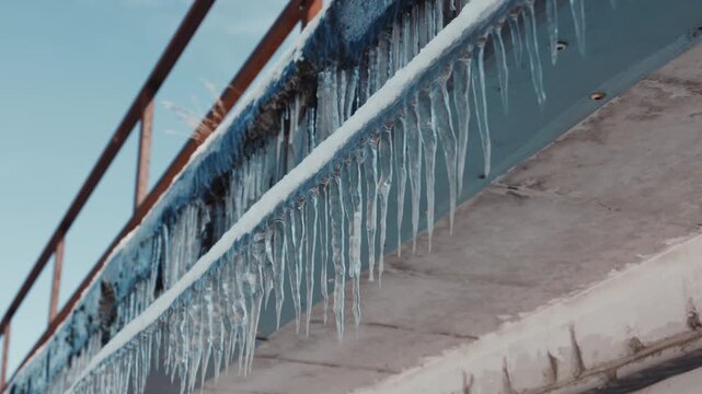 A low angle view shows sharp icicles hanging from a concrete roof edge against a blue sky. Concept of freezing temperatures, harsh winter weather conditions, and seasonal hazards.