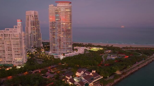 Aerial drone footage of Miami South Pointe Park at dusk captures luxury highrise condominiums with illuminated windows, lush tropical greenery, sandy beachfront, and calm Atlantic Ocean waters.