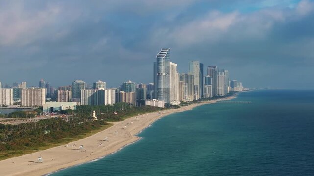 Aerial footage of Haulover Park in Miami, Florida showcasing sandy beaches alongside turquoise Atlantic waters. Luxury high-rise condominiums and skyscrapers line coastline under dramatic cloudy.
