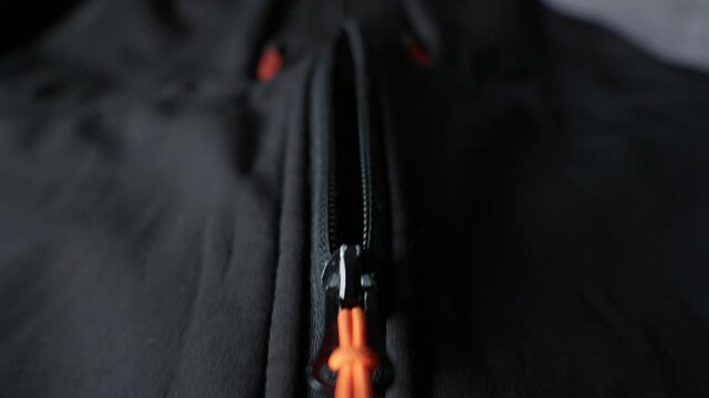 A macro close-up highlights a black zipper featuring a bright orange pull cord on dark fabric. Concept of outdoor gear design, secure fastening, and modern clothing manufacturing.