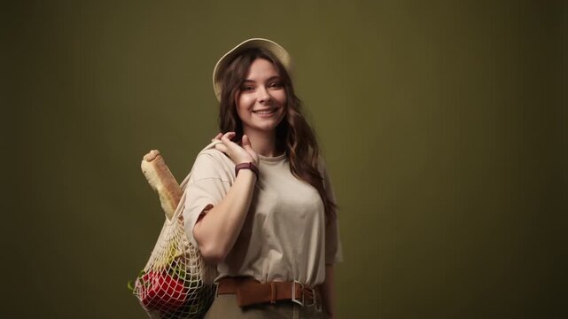 Cheerful woman carrying reusable mesh bag with fresh vegetables, fruits and bread walking and smiling