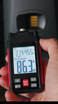A person holds a digital sound level meter that displays a noise reading of eighty-three decibels. The device also shows temperature and humidity readings in a dark studio room.