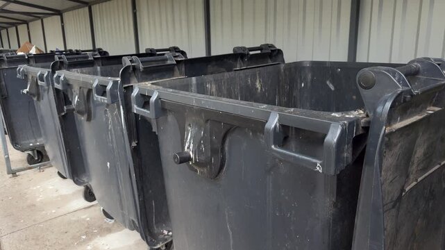 Row of empty black industrial dumpsters with wheels lined up under a covered metal structure. waste containers, recycling bins, commercial, industrial, outdoors