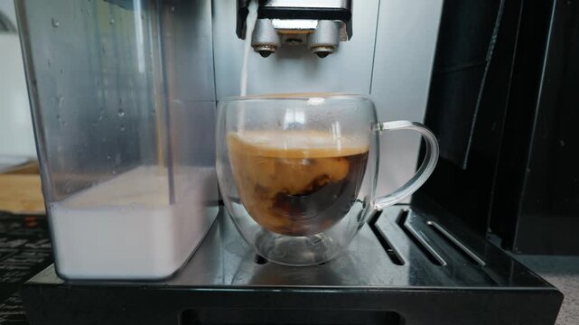 A modern espresso machine pours hot milk into a clear glass cup filled with dark coffee. Concept of morning routines, modern home appliances, and daily caffeine preparation.