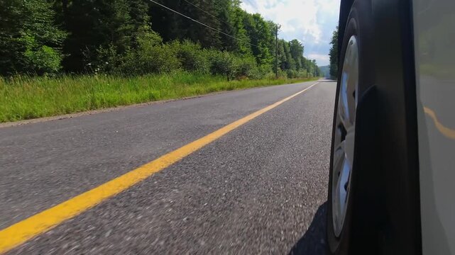 Rural countryside highway asphalt road drive. Cinematic POV of the front left car wheel driving plate. Yellow line lane separator in the frame. Cottage country commute, back of wheel exterior view.