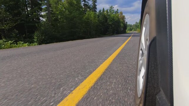 Rural countryside highway asphalt road drive. Cinematic POV of the front left car wheel driving plate. Yellow line lane separator in the frame. Cottage country commute, back of wheel exterior view.
