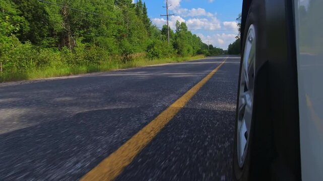Rural countryside highway asphalt road drive. Cinematic POV of the front left car wheel driving plate. Yellow line lane separator in the frame. Cottage country commute, back of wheel exterior view.
