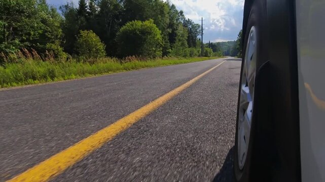 Rural countryside highway asphalt road drive. Cinematic POV of the front left car wheel driving plate. Yellow line lane separator in the frame. Cottage country commute, back of wheel exterior view.