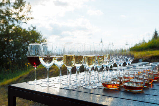 Rows of champagne flutes and whiskey glasses on a wooden table at an outdoor party.