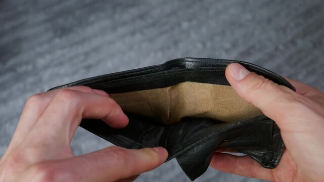 A man hands open an empty black leather wallet to show it contains no cash or cards. Concept of poverty, financial crisis, bankruptcy, inflation, and personal debt.