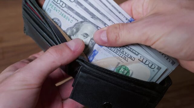 Hands carefully handle a thick stack of United States one hundred dollar bills inside a black leather wallet. Detailed view shows the texture of the currency against a dark wooden background