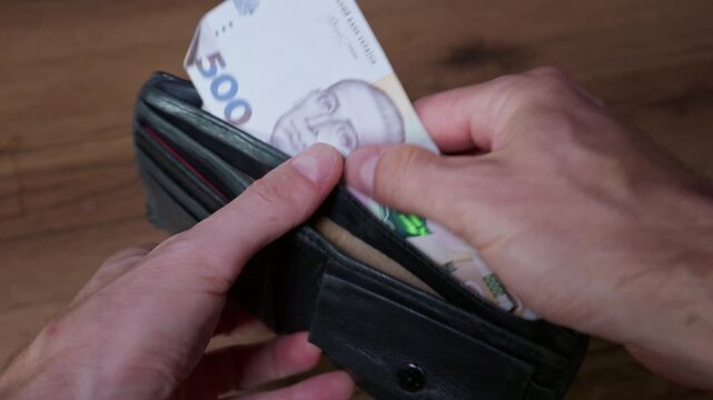 Close-up of male hands holding an open black leather wallet containing five hundred Ukrainian hryvnia banknotes. The person is preparing to pay with cash over a wooden background.