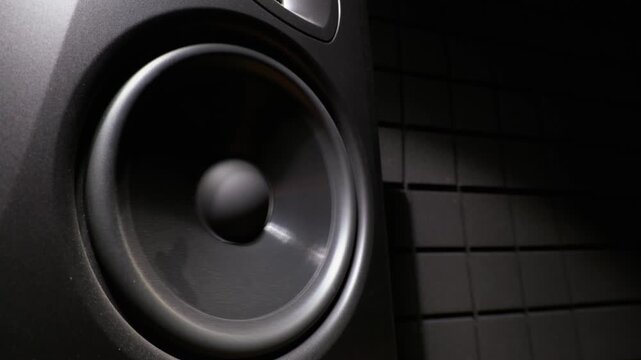 A professional studio monitor speaker cone is mounted in a dark soundproof room. Textured black acoustic foam panels line the wall in the background under dramatic side studio lighting.