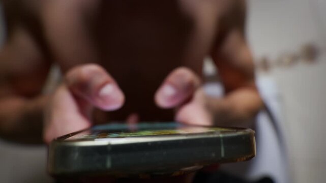 An unrecognizable man scrolls through social media apps on his smartphone while sitting on a toilet. The focus is on the mobile device and hands with a blurred background showing a bare chest