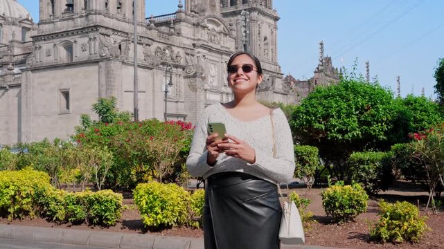 Young smiling Latina woman feeling happy, relaxed while typing and checking her smartphone, then looking up and enjoying the moment in Mexico City near a historic cathedral, wearing casual chic outfit