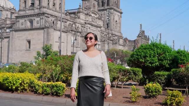 Young smiling Latina woman feeling joyful, relaxed and confident while walking and looking around, enjoying a sunny day in Mexico City near a historic cathedral, wearing casual chic outfit
