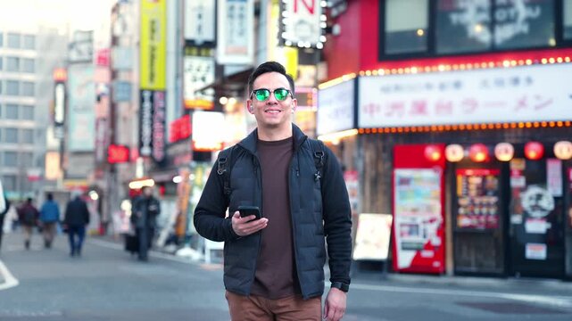 Young smiling Latino man feeling happy, relaxed while walking and checking his smartphone in Akihabara Tokyo Japan, surrounded by electronics shops colorful signage, wearing casual outfit sunglasses