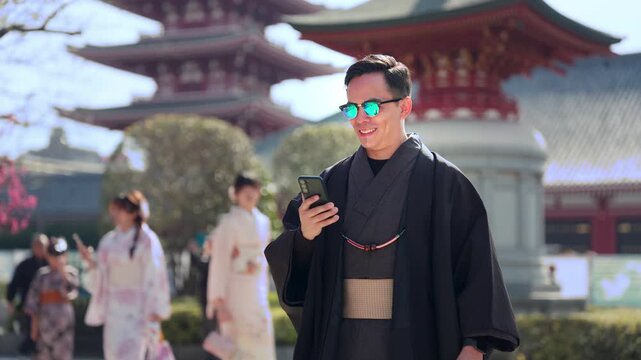 Young smiling Latino man feeling happy, curious and relaxed using his smartphone at Sensoji Temple in Asakusa Tokyo Japan, iconic historic site, traditional architecture, wearing kimono and sunglasses