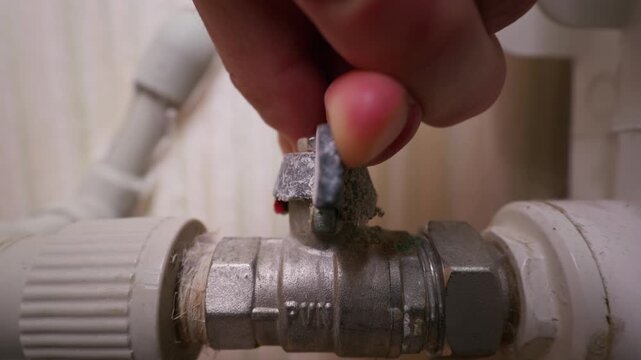Close-up view of a hand closing a limescale covered ball valve on a plumbing pipe in a home interior. Concept of plumbing maintenance, water conservation, home repair, and utility control.