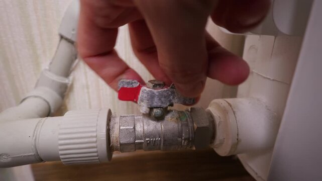 A hand turns a metal radiator valve on a household pipe to regulate the flow of hot water. Concept of energy conservation, home utility maintenance, heating control, and temperature regulation.