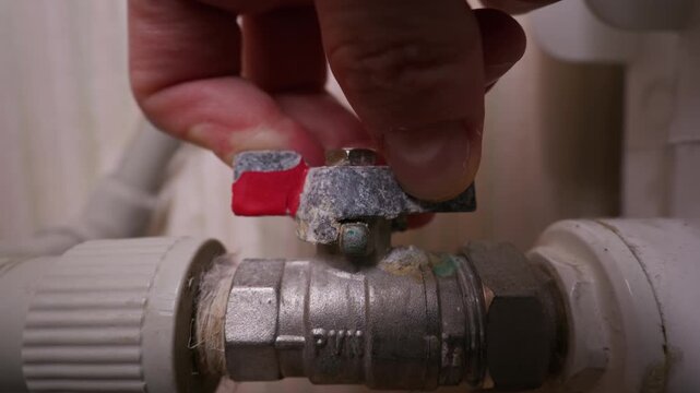 A close-up view shows a male hand turning a metallic ball valve on a polypropylene pipe. This crucial adjustment regulates the central heating system and water supply inside a residential home.