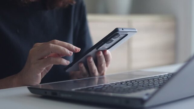 Woman sitting at living room and working on laptop and smartphone in office. Detail shot female hand messaging on smartphone before notebook. Concept of distant work freelance at home. Work from home.