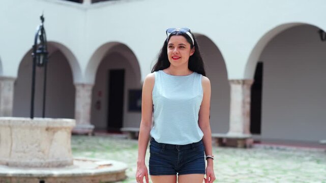 Young smiling Latina woman relaxed, happy and confident while walking at Universidad Autonoma de Yucatan in Merida Mexico, historic colonial courtyard, arches and fountain wearing casual summer outfit