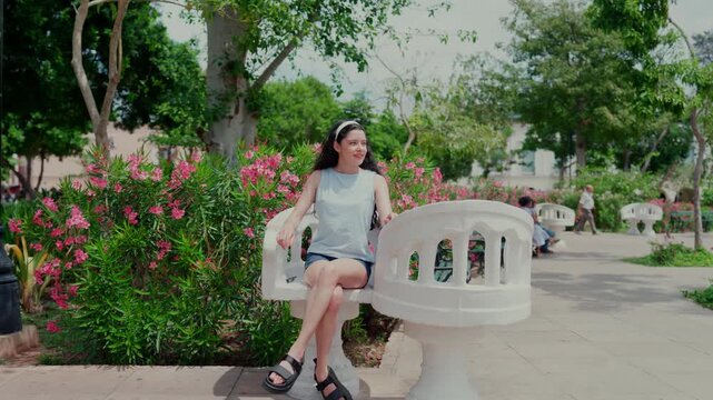 Young smiling Latina woman happy, calm and relaxed while sitting and looking around on famous Sillas Confidentes in Plaza Grande, Merida Yucatan Mexico, wearing striped top, denim shorts and sandals