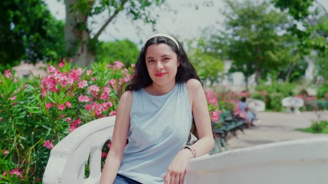 Young smiling Latina woman happy, calm and relaxed while sitting and looking around on famous Sillas Confidentes in Plaza Grande, Merida Yucatan Mexico, wearing striped top, denim shorts and sandals