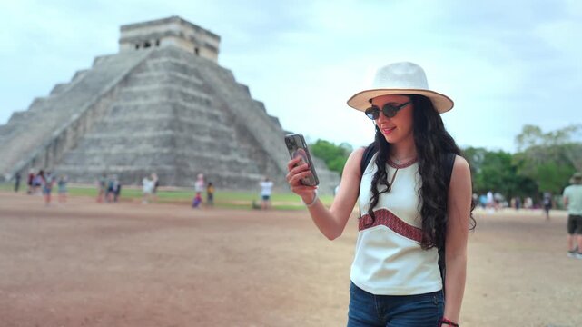 Young smiling Latina woman feels happy, curious while checking her smartphone at Chichen Itza in Yucatan Mexico, with Kukulcan pyramid ancient grounds behind her, wearing hat, sunglasses, casual top