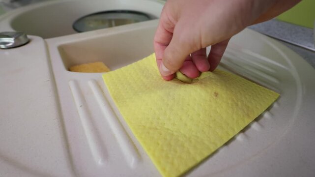 A person hand wipes the textured drainboard surface of a beige composite kitchen sink using a bright yellow cleaning cloth. The scene captures a common domestic chore in a modern home.