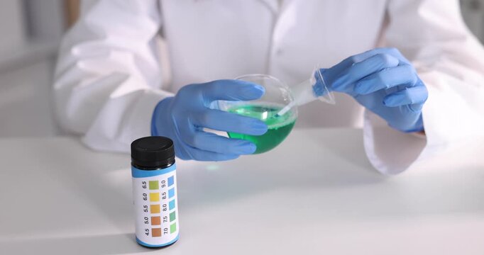 Scientist holding PH test strip and flask with liquid at white table in laboratory, closeup