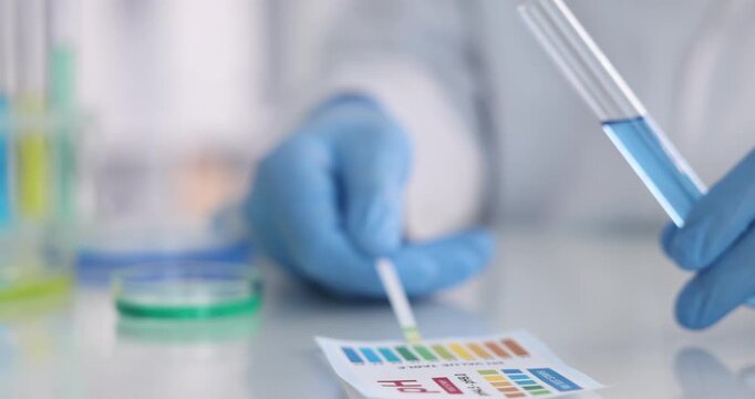Scientist holding PH test strip with color table and glass tube with liquid at white table in laboratory, closeup