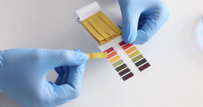 Scientist holding PH test strips with color table at white table in laboratory, closeup