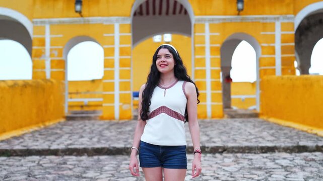 Young smiling Latina woman feels joyful, relaxed and confident while posing and looking at camera at the Convent of San Antonio de Padua in Izamal Yucatan Mexico, wearing white top with colorful trim