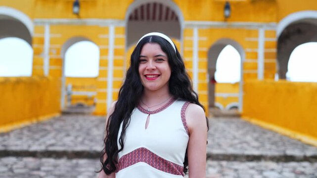 Young smiling Latina woman feels joyful, relaxed and confident while posing and looking at camera at the Convent of San Antonio de Padua in Izamal Yucatan Mexico, wearing white top with colorful trim
