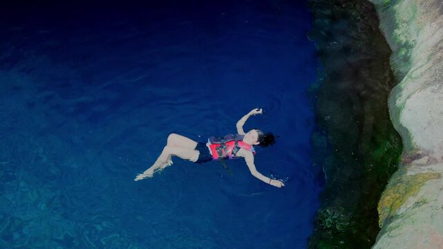 Young smiling Latina woman relaxed and adventurous while floating and swimming in a deep blue cenote in Yucatan Mexico, surrounded by clear freshwater and rocky walls, wearing life jacket and swimwear